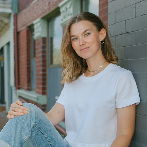 headshot of Mimi Millard, a white woman with dark blonde hair wearing a white t shirt and blue jeans, leaning against a brick wall.